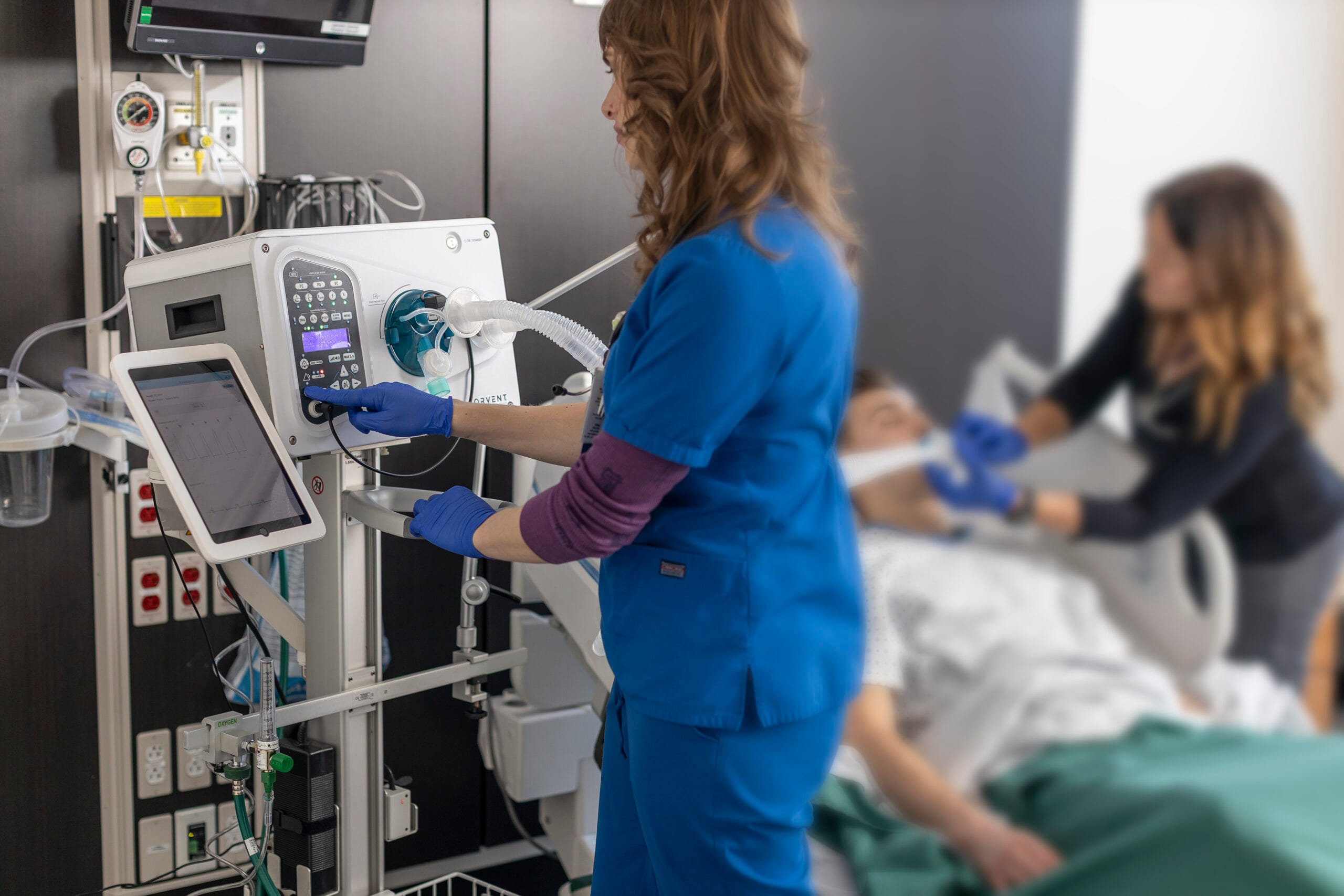 Two healthcare professionals assist a male patient on a hospital bed using a CorVent Medical ventilator. One clinician in blue scrubs adjusts settings on the ventilator, while the other in black checks the patient's non-invasive ventilation mask. A digital tablet displays patient data, and the ventilator is connected to wall-mounted medical gas and power outlets.